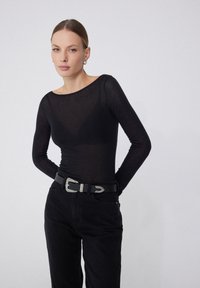 Haut long à manches transparentes noir avec un col rond associé à un jean noir taille haute et une ceinture décorative avec une boucle en argent.