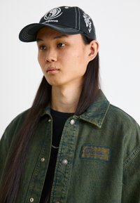 Jachetă din denim verde cu broderie albastră, nasturi prindere argintii, tricou negru dedesubt și o șapcă neagră cu modele și logo.
