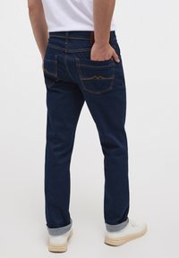 Jeans din denim de culoare albastru închis, cu croi drept, având două buzunare pe spate cu cusături portocalii și un tiv răsucit. Purtați cu adidași albi.
