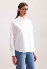 Camicia bianca con colletto aperto, maniche lunghe e orlo curvo. Indossata con jeans in denim azzurro chiaro. Tessuto dalla consistenza morbida.