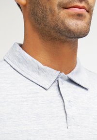 Polo shirt gris en tissu texturé, avec un col, deux boutons et de petits détails en fils bleu clair.