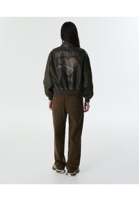 Chaqueta bomber verde olivo con parches de codo acolchados y gráfico de "AMOR FASCINANTE" en diseño de corazón rojo en la parte posterior, combinada con pantalones marrones.