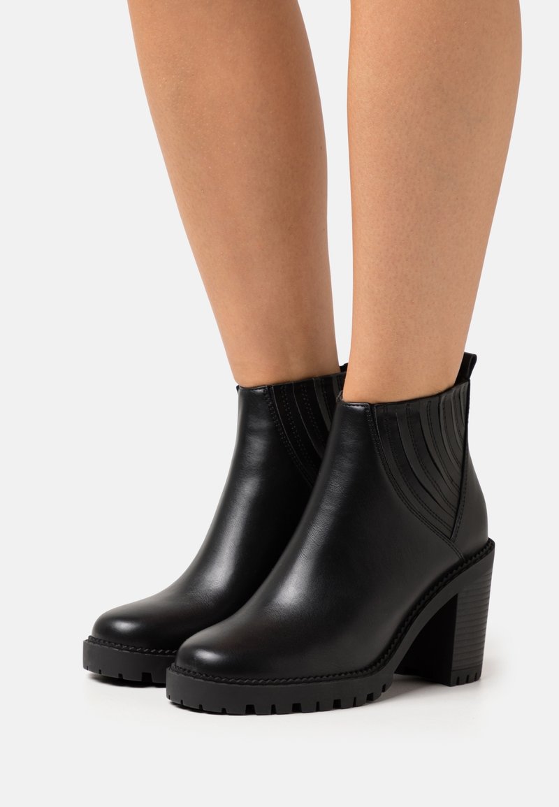 Bottines en cuir noir avec talons carrés et panneaux latéraux élastiques portées sur jambes nues sur un fond uni.