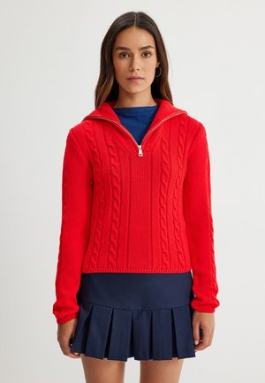 Roter Kabelstrick-Pullover mit halbem Reißverschlusskragen, langen Ärmeln und gerippten Bündchen. Kombiniert mit einem dunklen plissierten Rock.