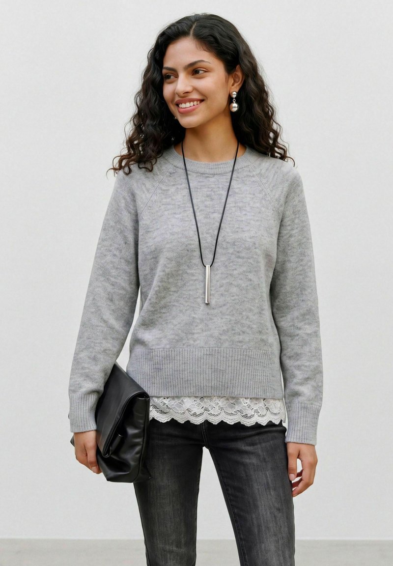 Jeune femme aux cheveux bouclés portant un pull gris, un jean noir, tenant une pochette noire et souriant sur un fond uni.