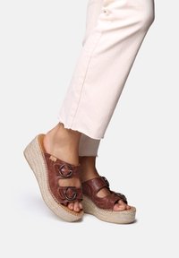 Sandali con zeppa marroni, dotati di due cinturini regolabili, design a fiori in rilievo e piattaforma in juta intrecciata; pantaloni beige chiaro con orlo sfrangiato.