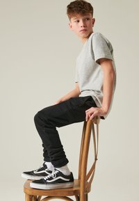 Tricou gri cu mâneci scurte, pantaloni de trening negri și adidași negri cu accente albe. Modelul stă pe un scaun de lemn, cu fața ușor întoarsă.