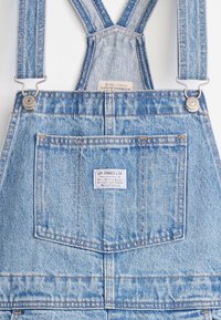 Ανοιχτό γαλάζιο τζιν σακάκι με ρυθμιζόμενους ιμάντες, μπροστινή τσέπη και ταμπελάκι πίσω με λευκό και μπλε κείμενο. Κλασικός σχεδιασμός σακακιού.