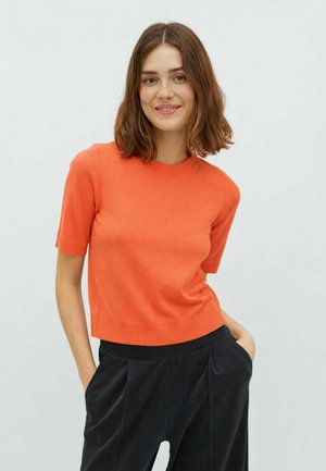 Femme aux cheveux bruns mi-longs portant un pull orange à manches courtes et un pantalon noir, souriant avec les mains dans les poches.