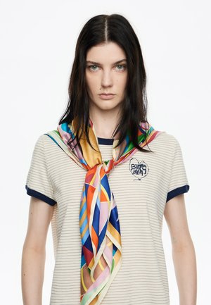 Camiseta de rayas beige con ribete azul marino, que presenta un logo. Una colorida bufanda de seda con patrones geométricos está enrollada alrededor del cuello.