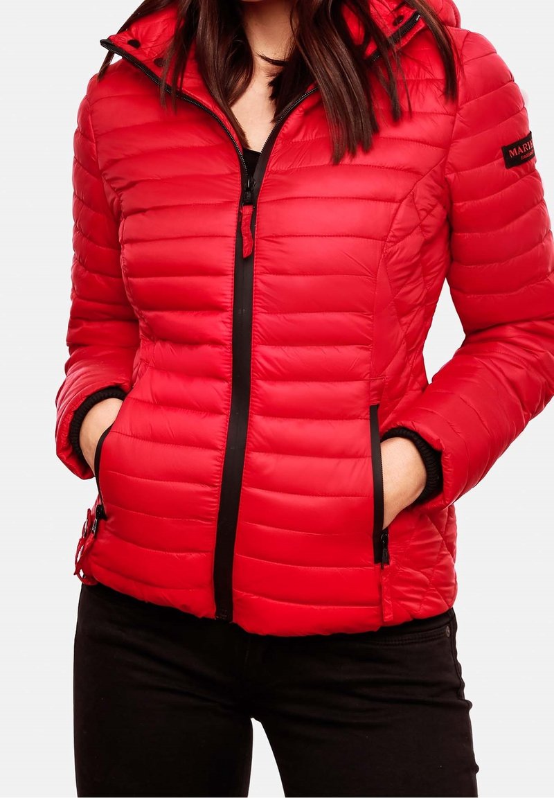 Frau in einer roten gesteppte Jacke mit Kapuze, schwarzem Reißverschluss und Händen in den Seitentaschen, kombiniert mit schwarzen Hosen.