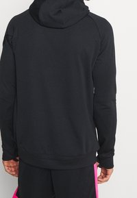 Osoba ubrana w czarną bluzę z kapturem i czarne szorty z jasnoróżowymi bocznymi paskami, pokazana od tyłu.