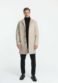 Bežový vlnený kabát s klopovým golierom, zapínanie na predné gombíky a bočné vrecká, nosený cez čierny rolák a čierne nohavice.