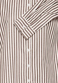 Brun og creme-stribet skjorte med knapper, der har en nedfaldsfar, lange ærmer og hvide knapper langs placket og manchetter.