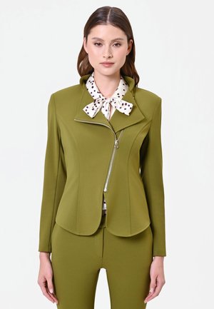 Femme portant une veste cintrée vert olive avec fermeture éclair asymétrique sur un chemisier blanc avec col nœud papillon à pois noirs, debout devant un fond uni.