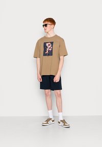 Beige oversized t-shirt with graphic print, paired with navy shorts and brown sneakers. Accessories include black sunglasses.