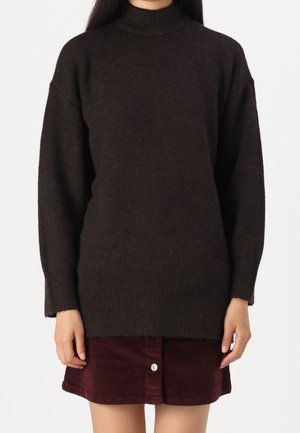 Kvinde iført en mørkebrun oversized højhalset sweater kombineret med en vinrød fløjlsmini nederdel med knapper foran.