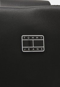 Un matériau noir texturé présente une étiquette rectangulaire argentée avec "TOMMY" et "JEANS" en lettres blanches sur fond noir.