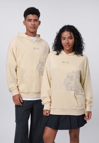 Felpa crema in misto cotone, con tasca frontale, vestibilità ampia e motivo grigio decorativo sul lato inferiore. Maniche e cappuccio semplici.