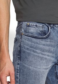 Jasnoniebieskie dżinsy o slim fit, z złotymi nitami i szczegółowym wykończeniem na kieszeniach i szwach. Miękka tekstura i klasyczny design.