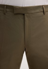 Nahaufnahme von olivgrünen Hosen mit verdecktem vorderem Knopfflug und Gürtelschlaufen vor weißem Hintergrund.