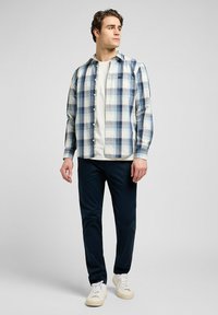Camicia a maniche lunghe a scacchi nei colori azzurro chiaro, blu scuro e bianco, abbinata a pantaloni blu navy e sneakers color crema. Uno stile casual con dettagli in bottoni.