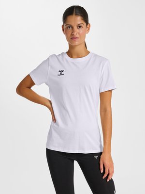 Жена стои с ръка на ханша, облечена в бяла тениска hummel и черен клин на чисто бял фон.
