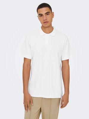 Noor mees lühikeste juustega, kandes tavalist valget lühikeste varrukatega polosärki ja beeže pükse, seismas hele taust.