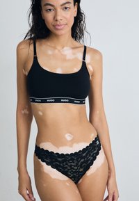 Brassière noire sans couture avec bande logo et bretelles réglables, associée à un string en dentelle noir avec un bord festonné et un motif floral.