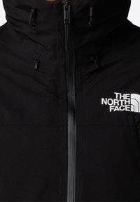 Chaqueta aislante negra de tela suave. Cuenta con un alto cuello, cremallera frontal y un logo blanco en la parte superior izquierda.