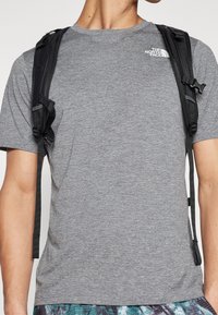 Šedé sportovní tričko s krátkým rukávem z texturované látky, s logem The North Face na hrudi a černými popruhy batohu.
