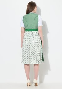 Corpiño a cuadros verdes con una falda blanca de estampado floral, que presenta pliegues y un lazo, combinada con tacones color crema. Vista trasera.