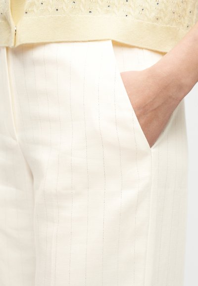 Το χέρι ενός ατόμου ακουμπάει μέσα στην τσέπη ενός παντελονιού εκρού με διακριτικές κάθετες ρίγες και ένα κίτρινο τοπ με μικρές διακοσμητικές λεπτομέρειες.
