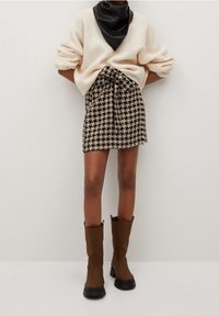 Maglione lavorato a maglia beige, sciarpa in seta nera al collo, gonna mini con motivo a pied de poule e vita con cravatta, e stivali di gomma marroni fino al ginocchio.