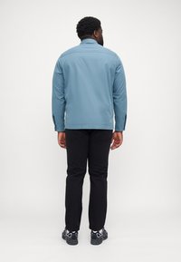 Giacca azzurro chiaro con colletto, maniche lunghe e polsini con bottoni a pressione, abbinata a pantaloni neri e scarpe da ginnastica nere. Vista posteriore.