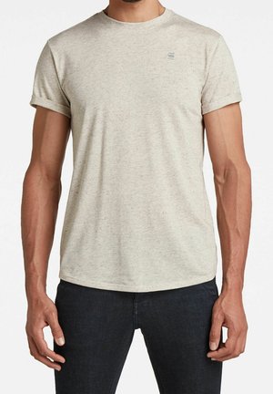 Mężczyzna w jasnobeżowej koszulce z krótkim rękawem, z podwiniętymi mankietami i małym logo na piersi, w połączeniu z ciemnoniebieskimi dżinsami.