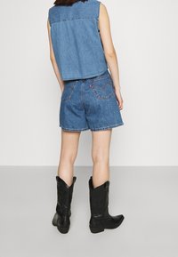Denimweste mit raw Saum, kombiniert mit blauen Denim-Shorts mit Gesäßtaschen. Schwarze Cowboystiefel mit kunstvollen Präge-details.