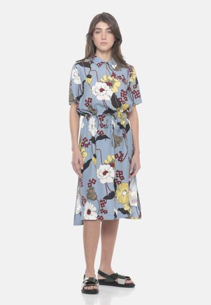 Donna che indossa un vestito midi blu a fiori con colletto, cintura in vita, maniche corte e sandali neri aperti davanti, su sfondo bianco neutro.