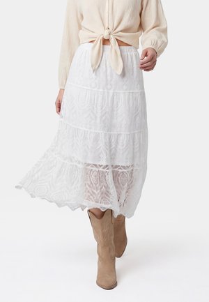 Frau trägt einen weißen Spitzen-Midirock, beige gebundene Bluse und braune Cowboy-Stiefel und geht auf neutralem Hintergrund nach vorne.