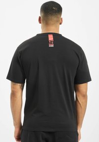 Black short-sleeve t-shirt with a round neckline, featuring a red tag on the upper back displaying a logo. Smooth, cotton-like texture.