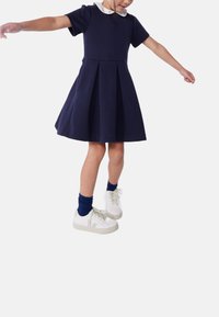 Σκούρο μπλε φόρεμα με κοντά μανίκια και πιέτες, λευκό γιακά, σετ με λευκά αθλητικά παπούτσια και σκούρες μπλε κάλτσες. Λιτή υφή και casual σχεδιασμός.