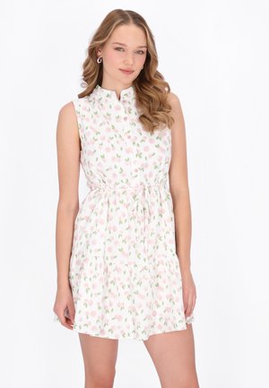 Fiatal nő hullámos hajjal, ujjatlan fehér ruhában, amely rózsaszín virágmintával és elöl gombokkal díszített, egyszerű háttér előtt áll.