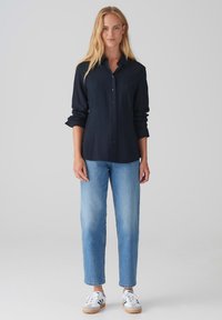 Camicia blu navy con bottoni e maniche lunghe, abbinata a jeans dritti azzurri chiari e sneakers bianche con dettagli neri.