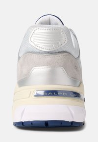 Šedé semišové a stříbrné syntetické tenisky s podpatkem a gumovou podrážkou; obsahují texturovaný stříbrný panel a branding "RALPH LAUR" v metalické modré.