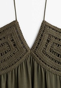 Gros plan sur un haut crocheté vert olive avec des motifs géométriques carrés et des fines bretelles sur un fond blanc.