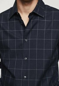 Uomo che indossa una camicia nera con bottoni, decorata da una griglia bianca, con colletto aperto e maniche corte visibili.