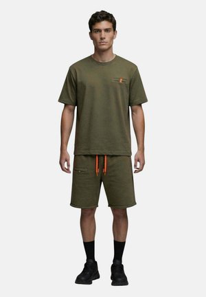 Homme debout face à l’avant portant un t-shirt vert olive et un short avec des cordons orange, des chaussettes noires et des chaussures de sport noires.