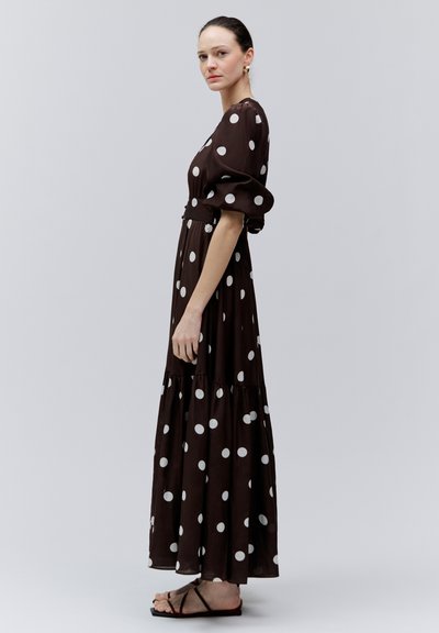 Mujer de perfil vestida con un largo vestido marrón oscuro con lunares blancos y mangas abullonadas, combinado con sandalias planas negras.