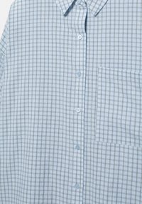 Vaaleansininen nappipaita, jossa on ohut harmaa ruutukuvio ja rintatasku oikealla puolella, kaulus näkyvissä yläosassa.