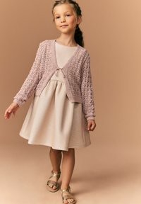 Cardigan tricotat roz deschis cu un design cu broderie din dantelă peste o rochie bej lucioasă. Rochia are o fustă evazată și o textură moale. Sandalele aurii completează ținuta.